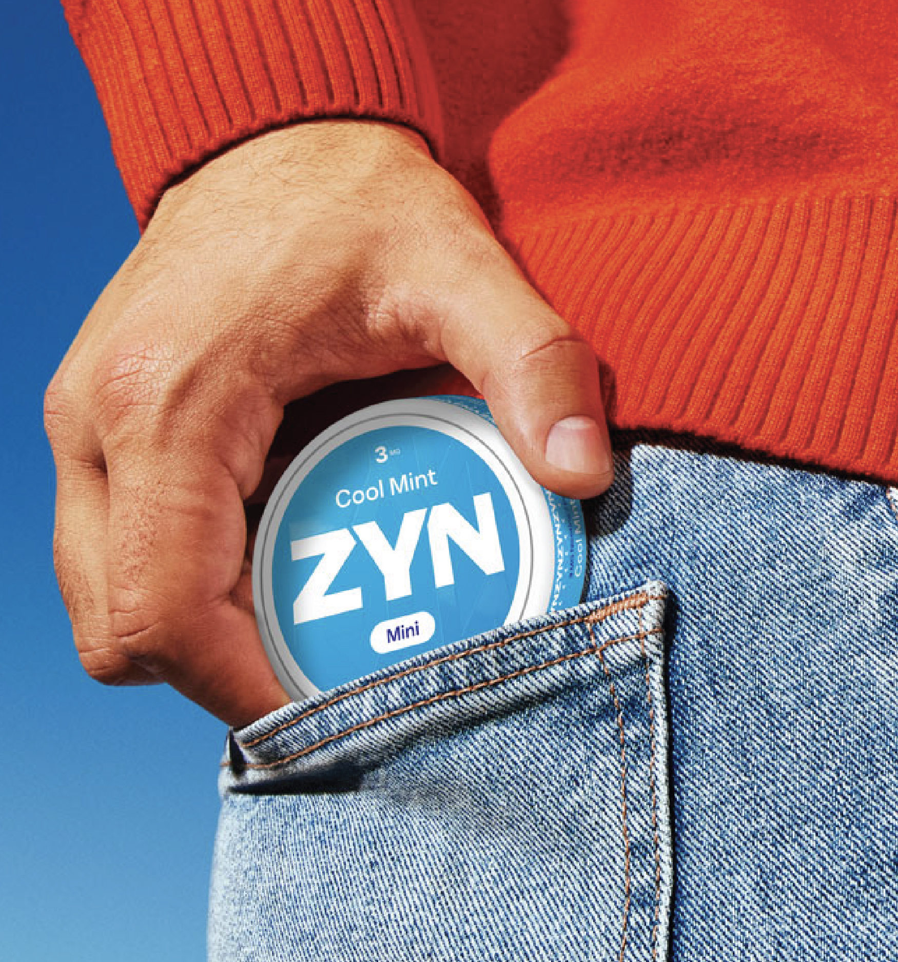 A person in a red sweater placing a red ZYN container into the back pocket of their blue jeans.